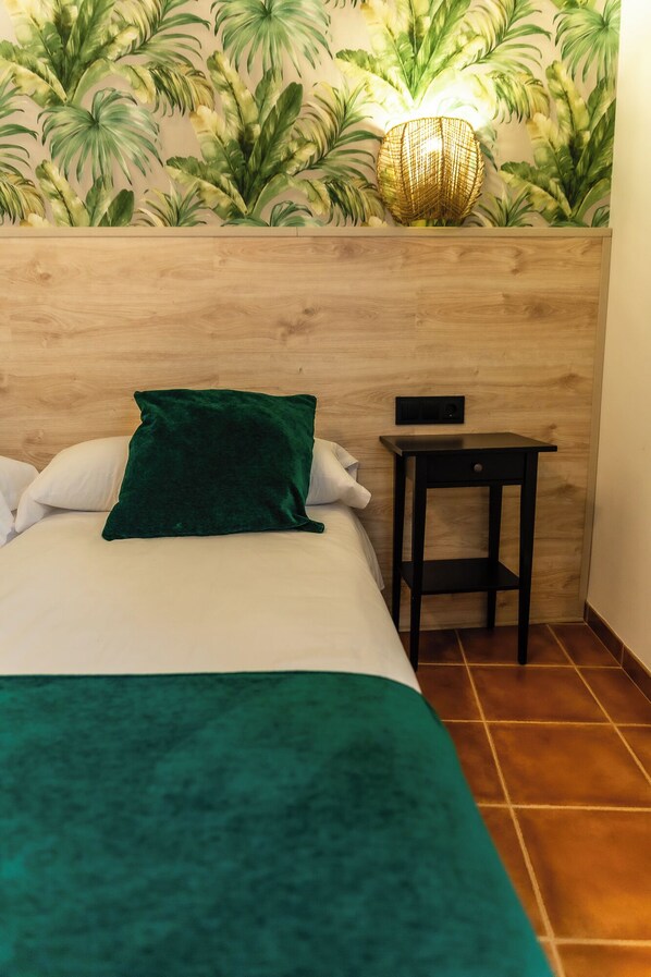 Habitación de hotel con cama, manta verde, cabecero de madera, una mesita y una pared con un mural de plantas.