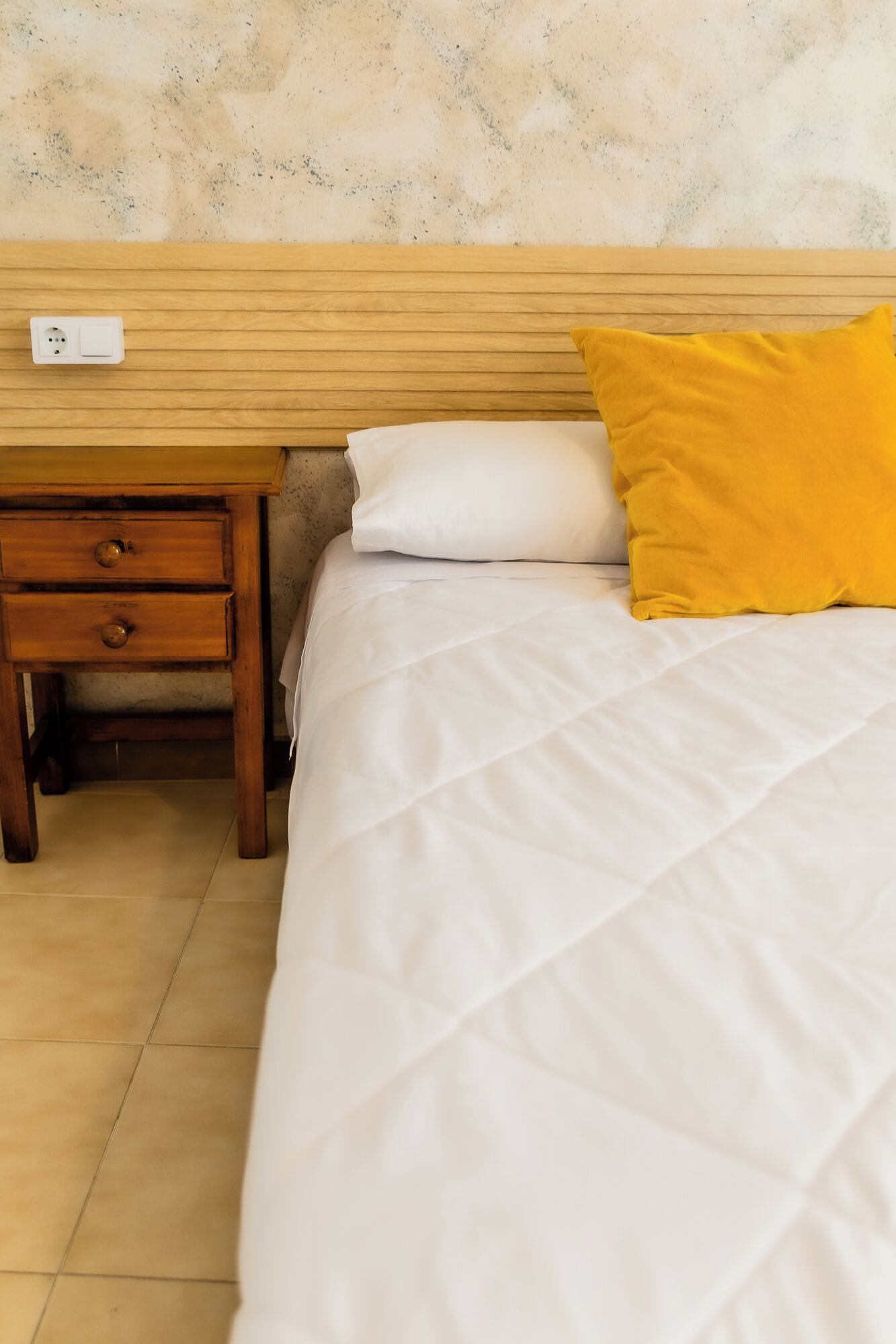 Ein Hotelzimmer mit einem hölzernen Kopfteil, einem weißen Bett mit einer gesteppten Decke, einem gelben Kissen und einem Nachttisch aus Holz mit Schubladen.