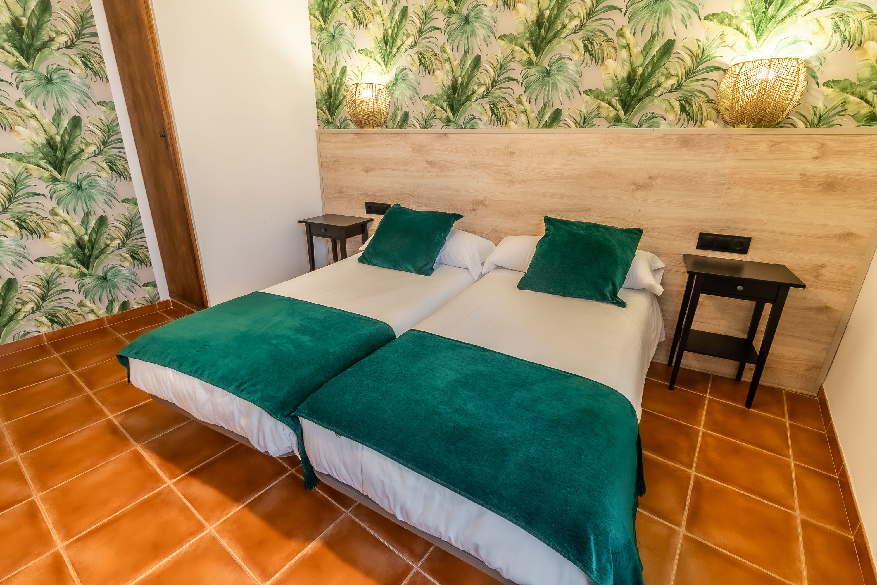 A double bed with green bedding, a wooden headboard, two bedside tables, and a wall with a tropical leaf wallpaper.