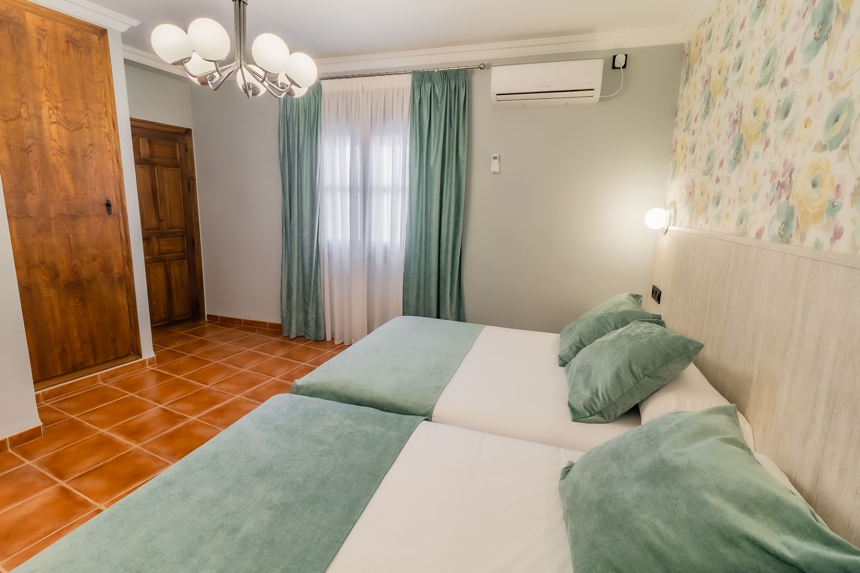 A room with two beds, a wooden door, a window with curtains, an air conditioner, and a light fixture.