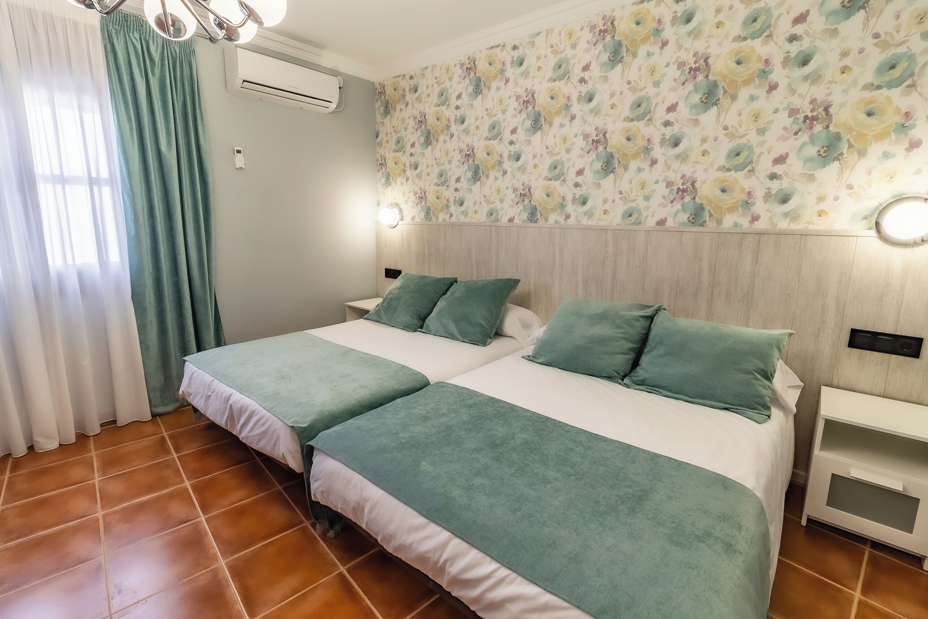 A double bed with green bedding, a nightstand, a wall-mounted air conditioner, and a floral-patterned wallpaper.