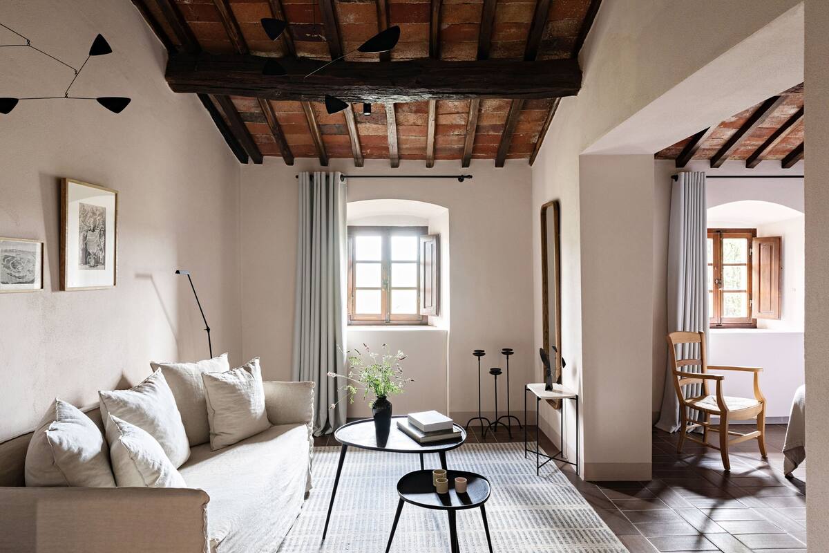 A cozy living room with a sofa, a small round table with a vase and books, and a wooden chair.