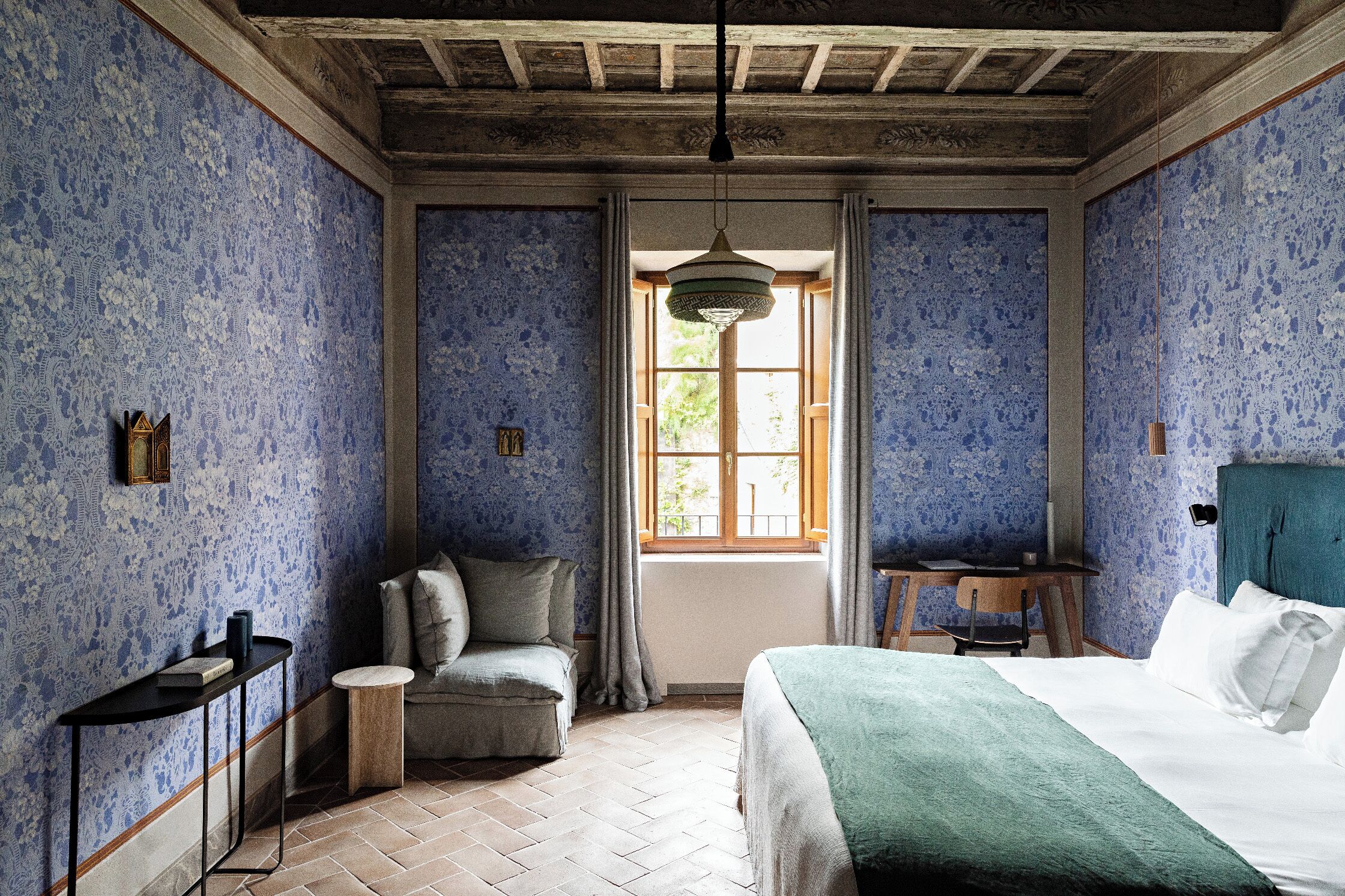 A room with a bed, a chair, a small table, and a window with curtains.