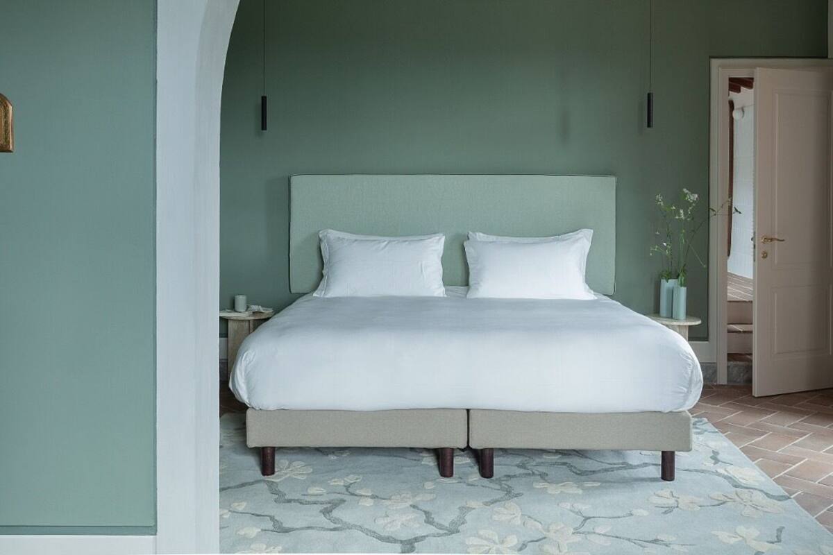 A bedroom with a large bed, a nightstand, a vase with flowers, and a door leading to another room.
