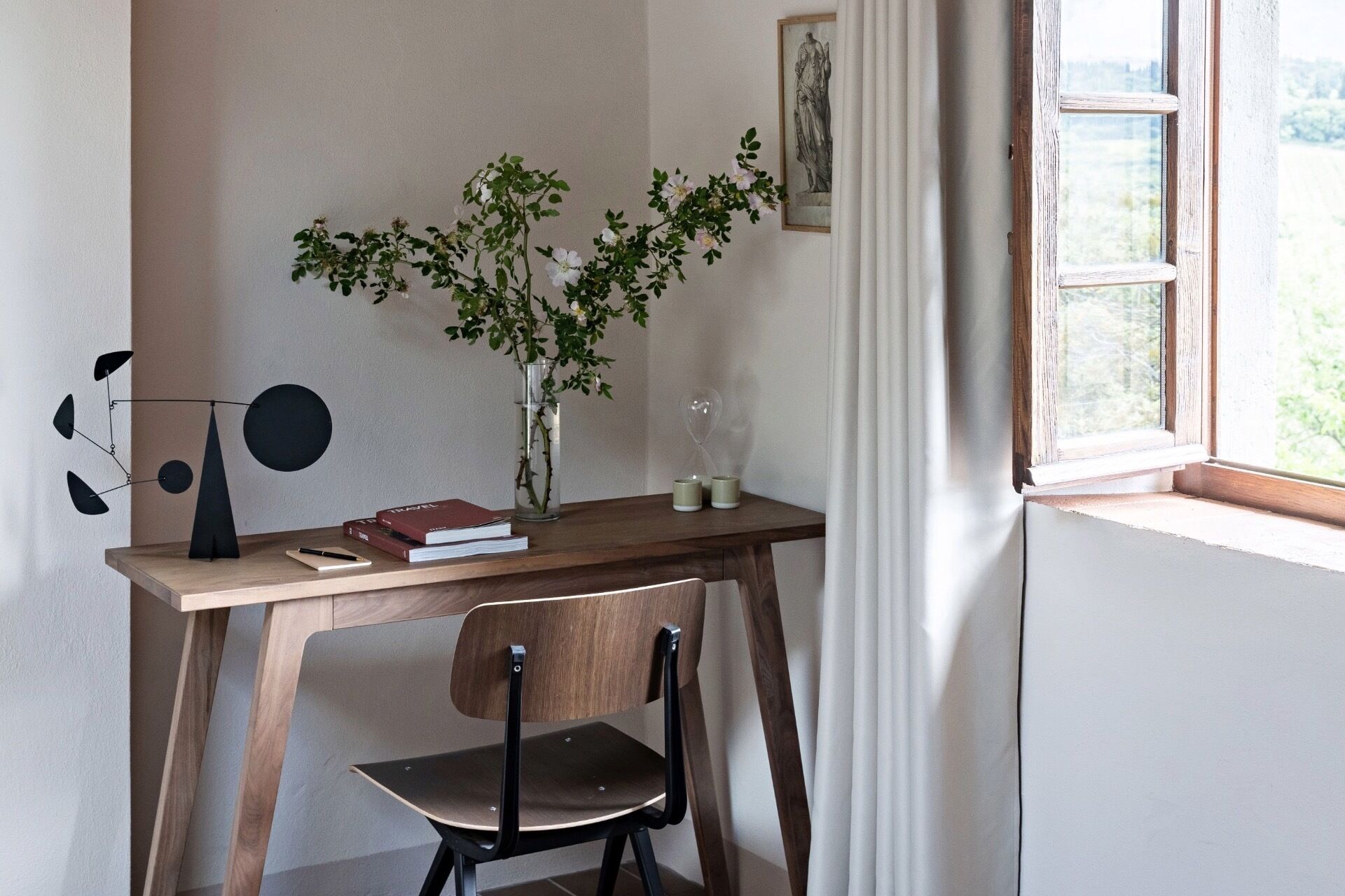 Un espacio de estudio con un escritorio de madera, una silla, un florero con flores, un reloj y una ventana con cortinas.