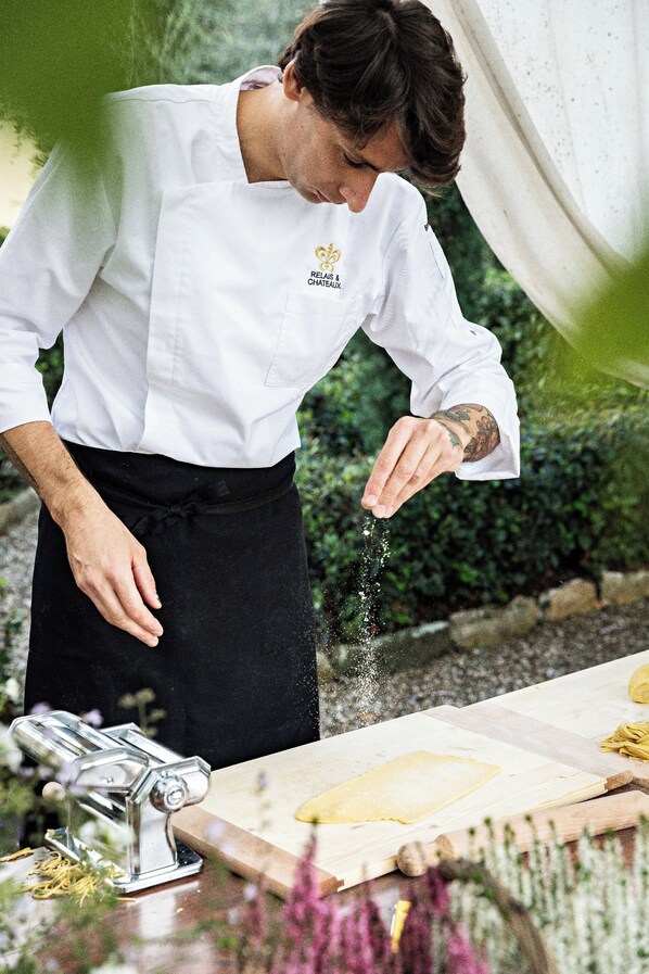 Una persona con bata de chef blanca, que lleva un logo, preparando comida en una mesa con una máquina para pasta y una tabla de cortar.