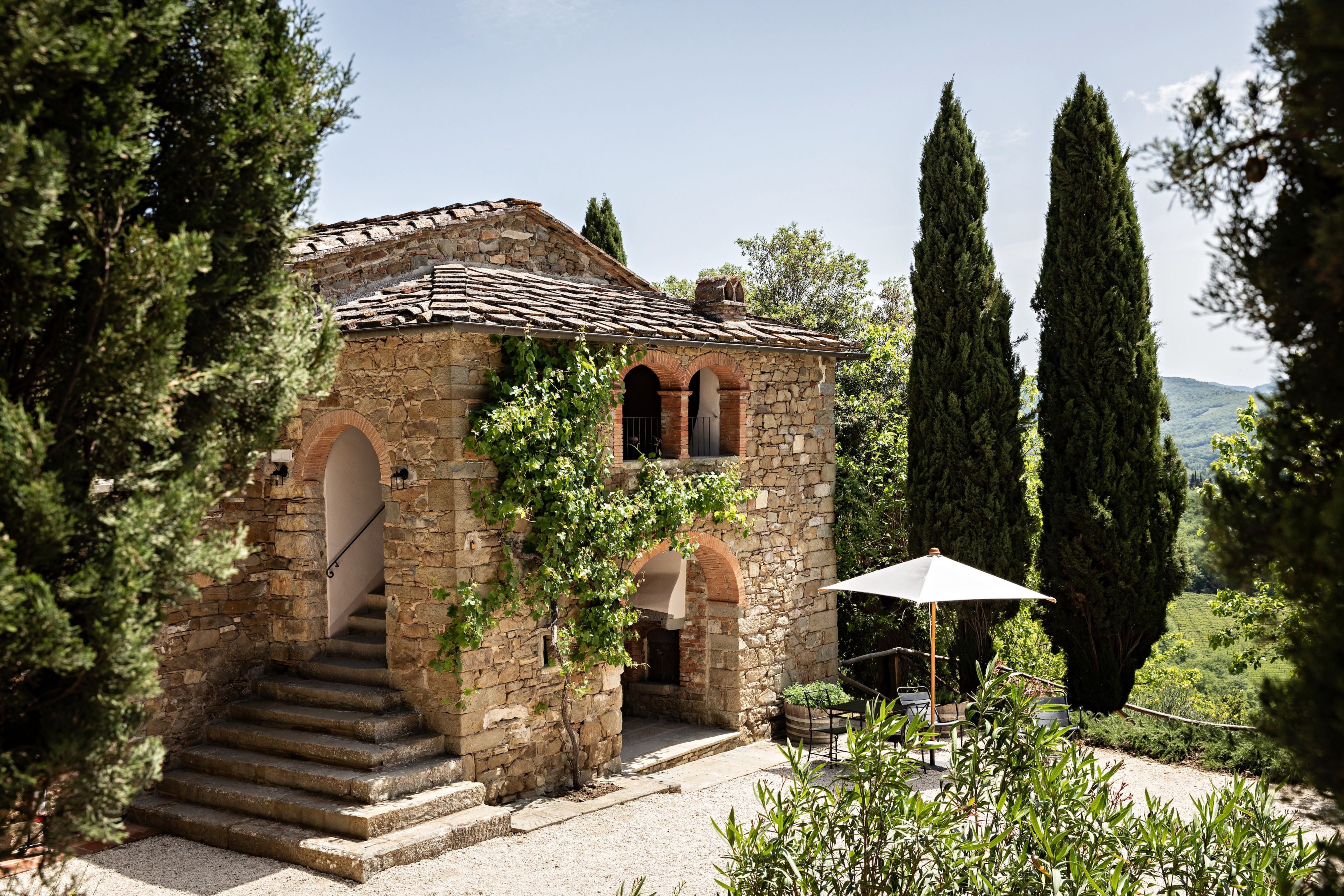 A stone building with a tiled roof, a staircase leading to an entrance, and a garden area with a table and chairs.