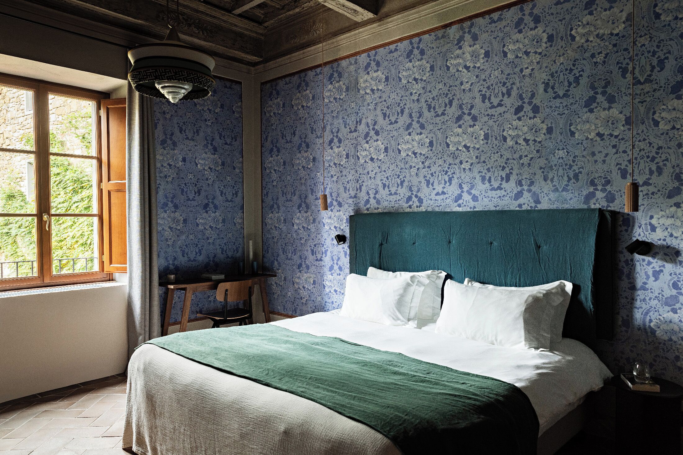 A bedroom with a large bed, a desk, and a window with a view of greenery.