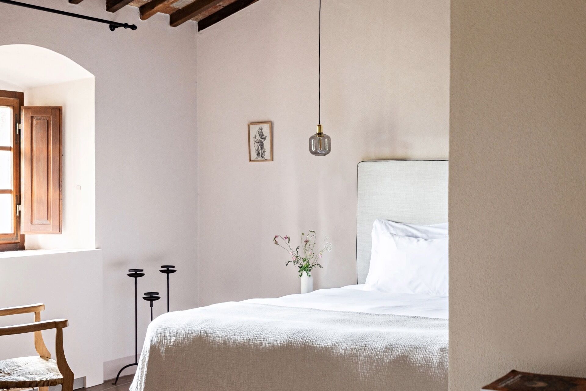 Une chambre à coucher comprenant un lit, une chaise, une fenêtre avec des volets en bois, un tableau accroché au mur, un petit vase avec des fleurs et un luminaire suspendu.
