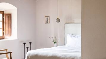 Une chambre à coucher comprenant un lit, une chaise, une fenêtre avec des volets en bois, un tableau accroché au mur, un petit vase avec des fleurs et un luminaire suspendu.