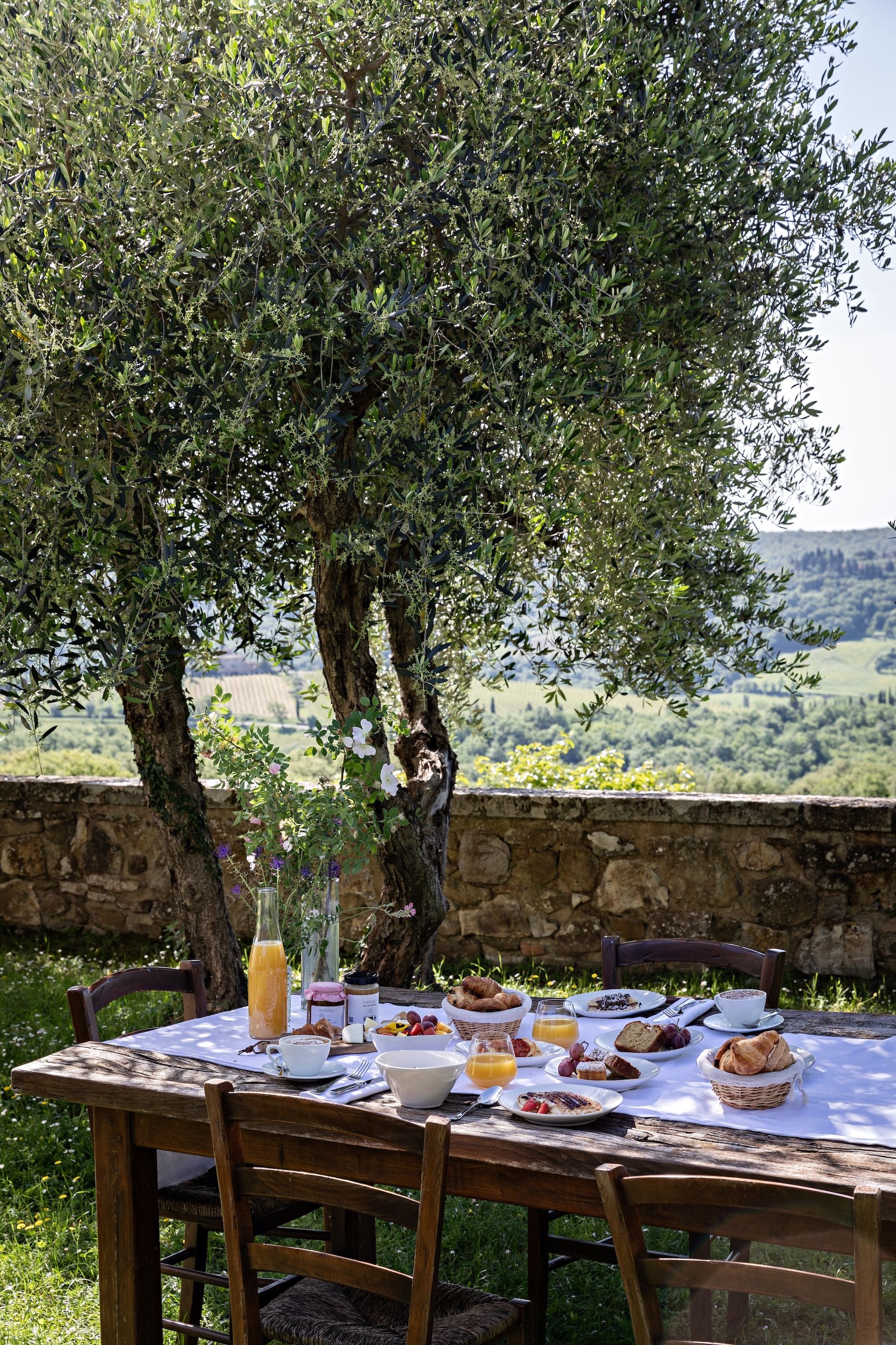 Une table à manger installée à l’extérieur, dressée pour le petit-déjeuner, avec une bouteille de jus d’orange et une vue sur un arbre et le paysage environnant.