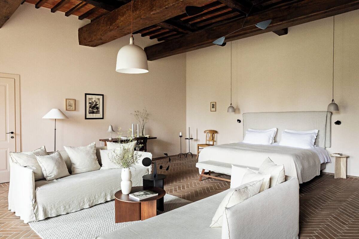 A bedroom with a large bed, a sofa, and a coffee table. There are two bedside tables and a lamp on each. The room has a herringbone floor pattern and wooden beams on the ceiling.