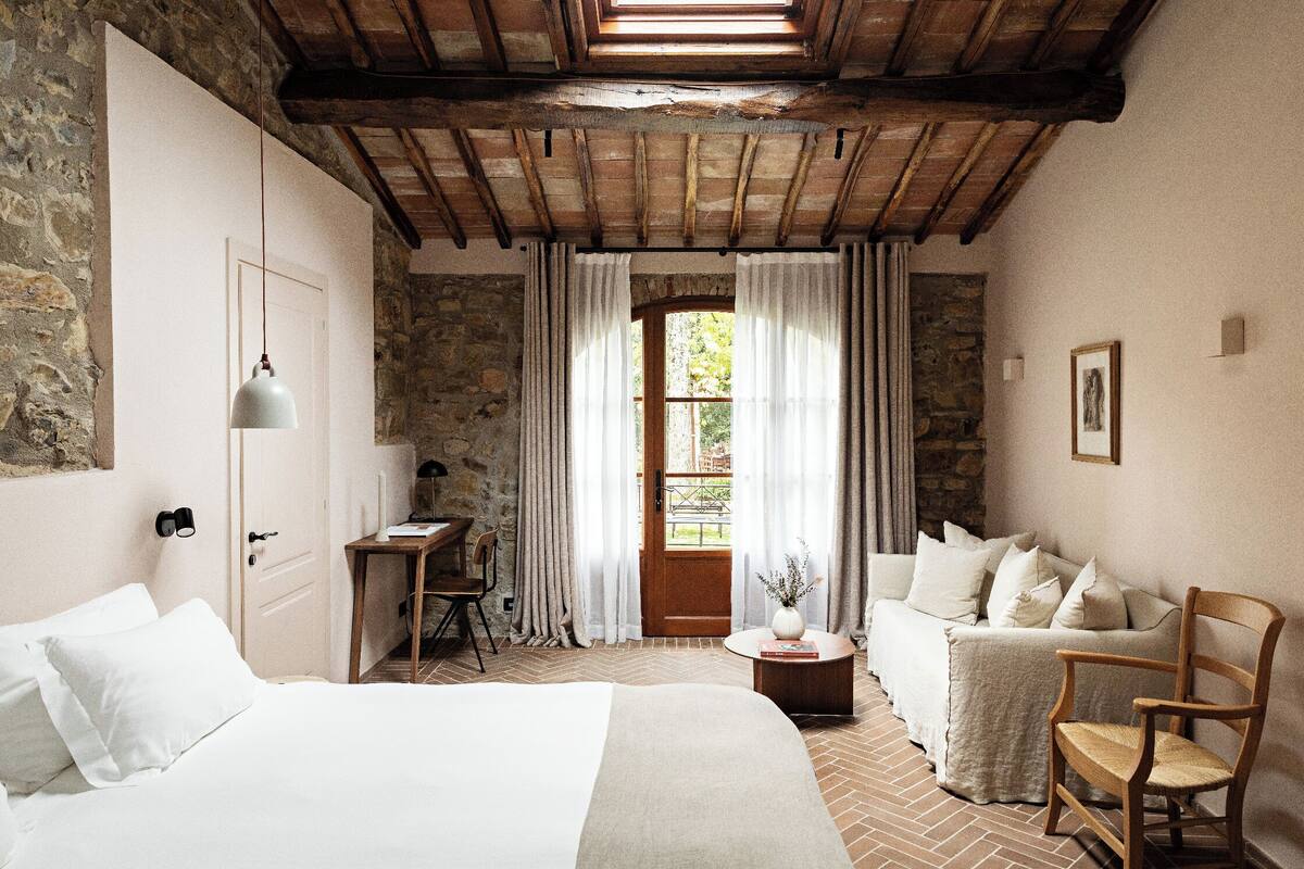 A bedroom with a wooden ceiling, a stone wall, a bed, a desk, a chair, a sofa, and a window with curtains.