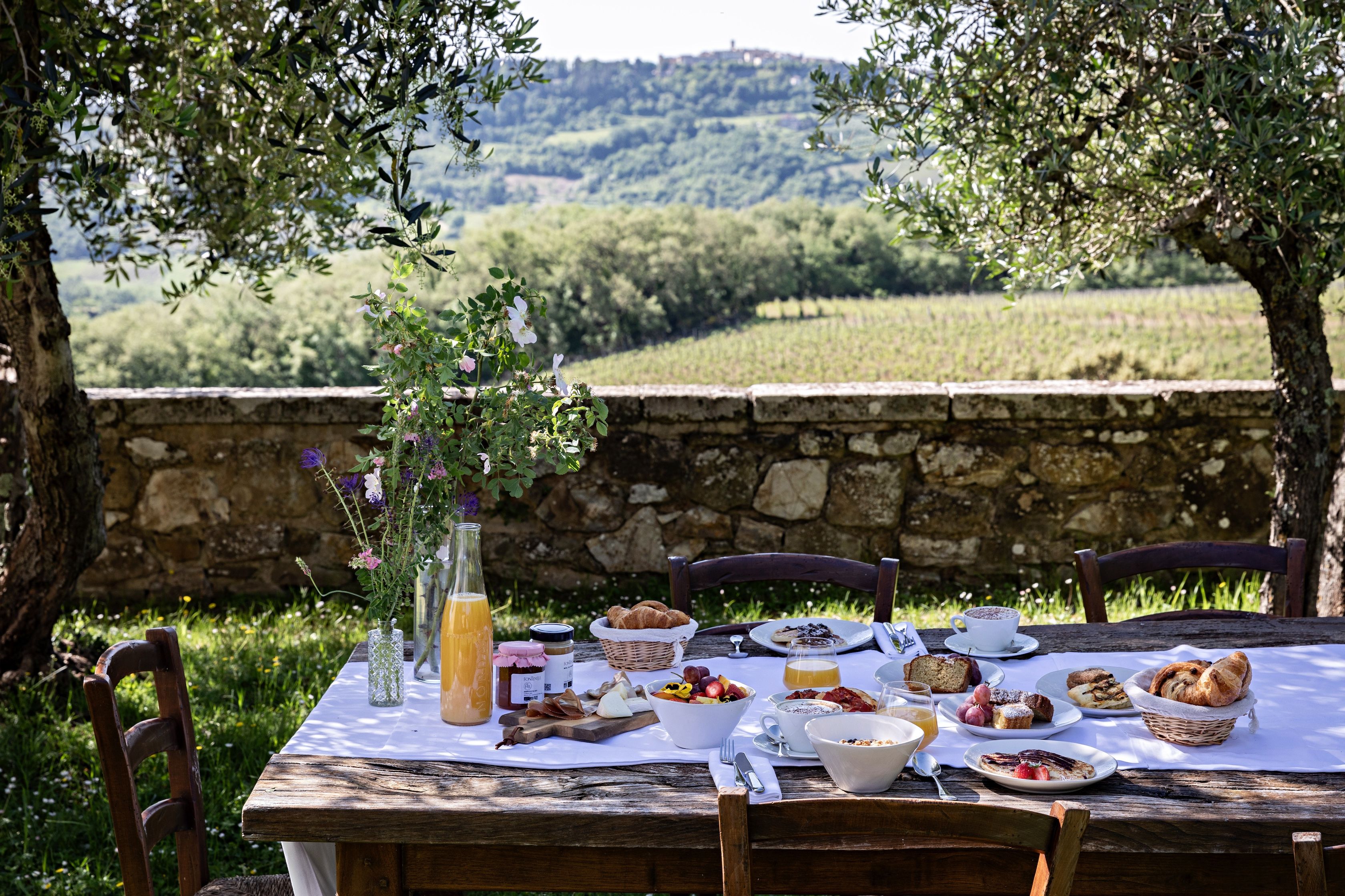 Mesa de comedor al aire libre con diversos alimentos y una botella de jugo de naranja, rodeada de muros de piedra y árboles.