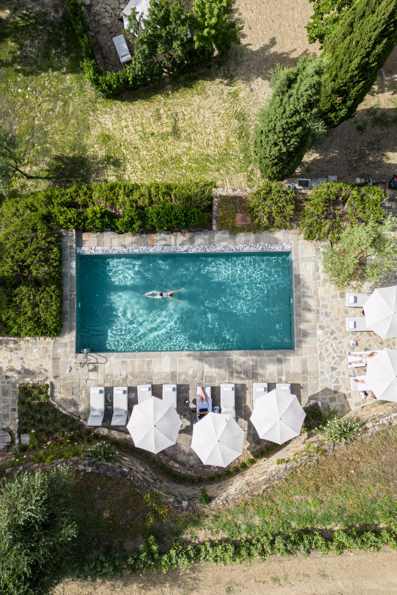 Eine Vogelperspektive zeigt einen rechteckigen Pool mit Steinrand, umgeben von Grünflächen und einer Steinterrasse mit weißen Sonnenschirmen.