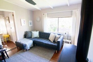 A living room with a sectional sofa, a small table, a lamp, and a window with curtains. - Banksia Cottage, pretty cottage with garden, pet freiendly (Leura)