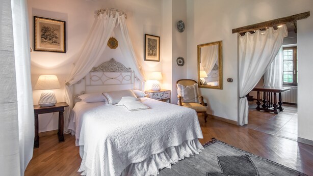 Une chambre à coucher avec un lit à baldaquin, des lampes de chevet, un fauteuil, un miroir et une vue sur l’extérieur.