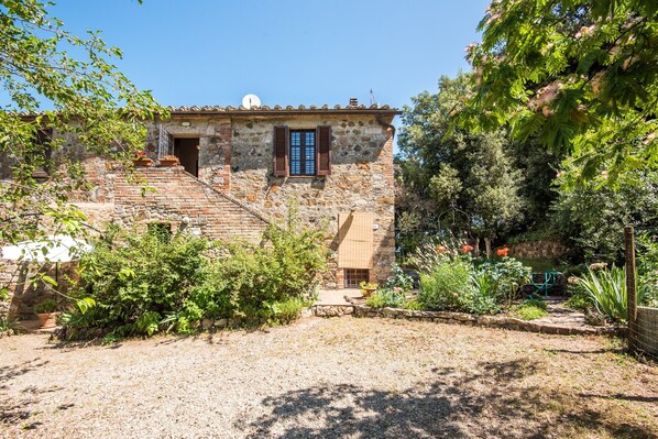 A rustic stone building with a small staircase, a window with shutters, and a garden area with various plants and a pathway.