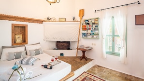 Een slaapkamer met een bed, een houten hoofdeinde, een kroonluchter, een open haard, een raam met gordijnen en een klein tafeltje met boeken.