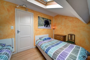 Ein Schlafzimmer mit schrägverdeckter Decke, einem Bett mit bunter Streifenoptik, einem Holznachttisch, einem kleinen Tisch, einer Wandleuchte und einer Tür.