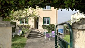 A building with a staircase leading to a door, a sign reading 'Cafes,' outdoor seating with chairs, and a green metal gate. - Luchon : maison flor (Bagnères-de-Luchon)