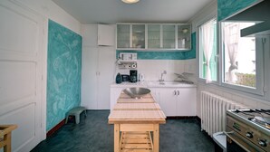 A compact kitchen with white cabinets, a wooden table, a sink, a stove, and a window with curtains. - Luchon : maison flor (Bagnères-de-Luchon)