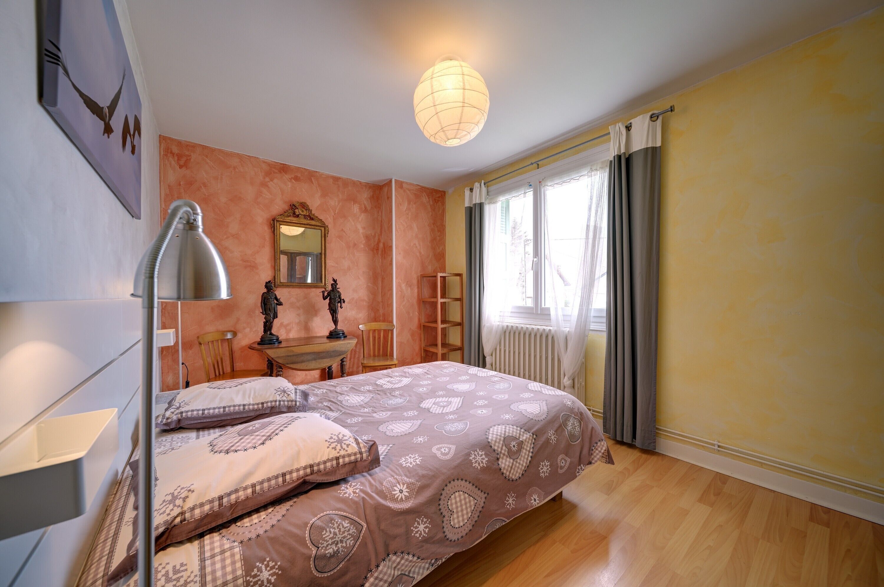 Een slaapkamer met een bed, een bureau, een stoel, een lamp, een spiegel en een raam met gordijnen.