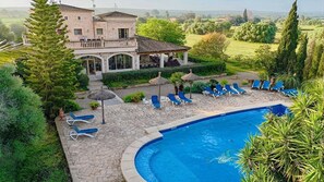 Un espace aménagé au bord de la piscine, avec des chaises longues, une terrasse en pierre et une grande maison dotée d’un balcon offrant une vue panoramique sur le paysage.