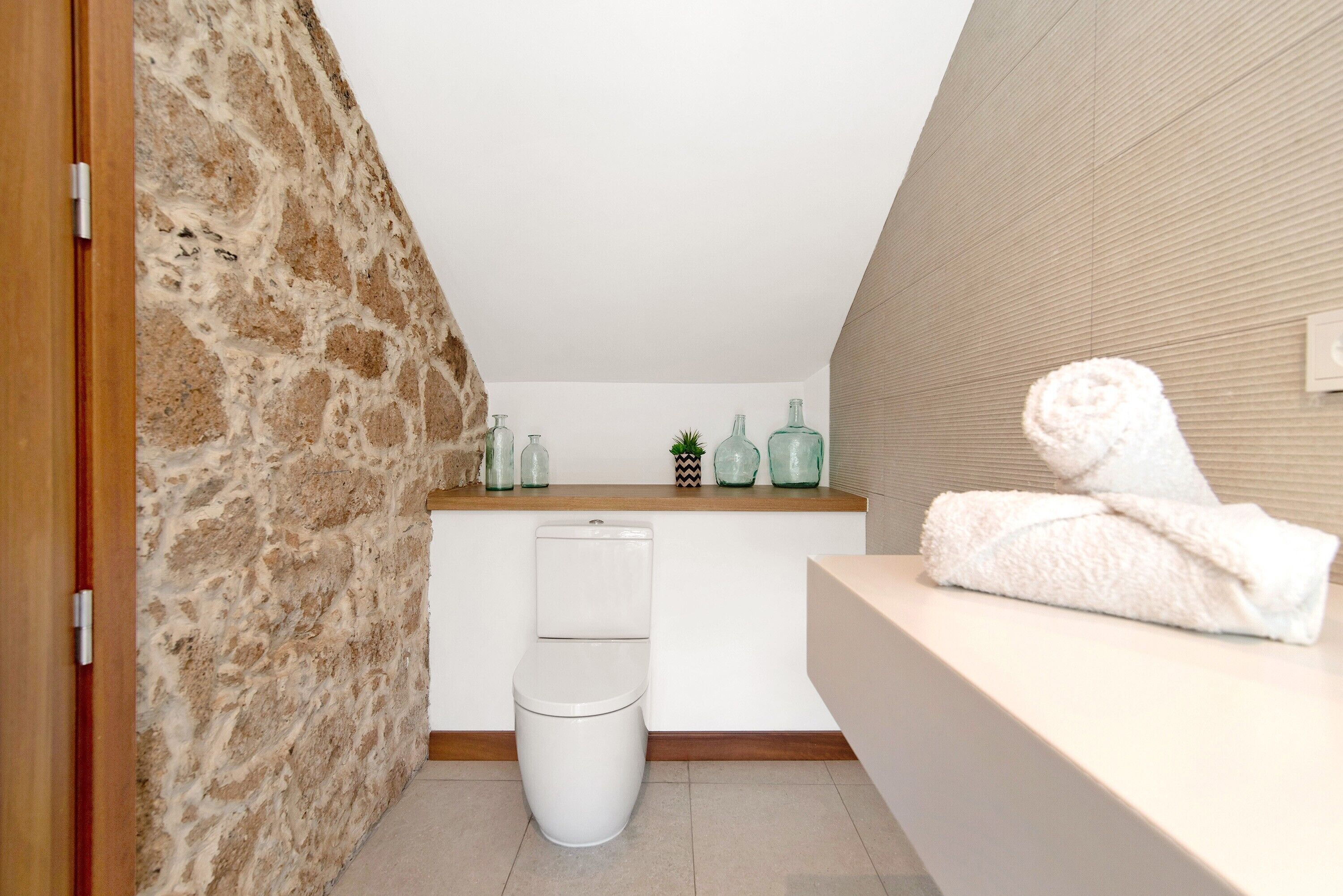 Un baño con pared de piedra, inodoro blanco, tocador de madera con botellas de vidrio y una toalla sobre la encimera.