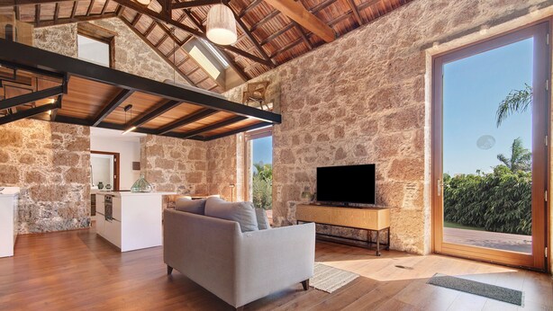 A spacious living area with a stone wall, wooden beams, a sofa, a TV, and a view of greenery.