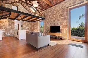 A spacious living area with a stone wall, wooden beams, a sofa, a TV, and a view of greenery.