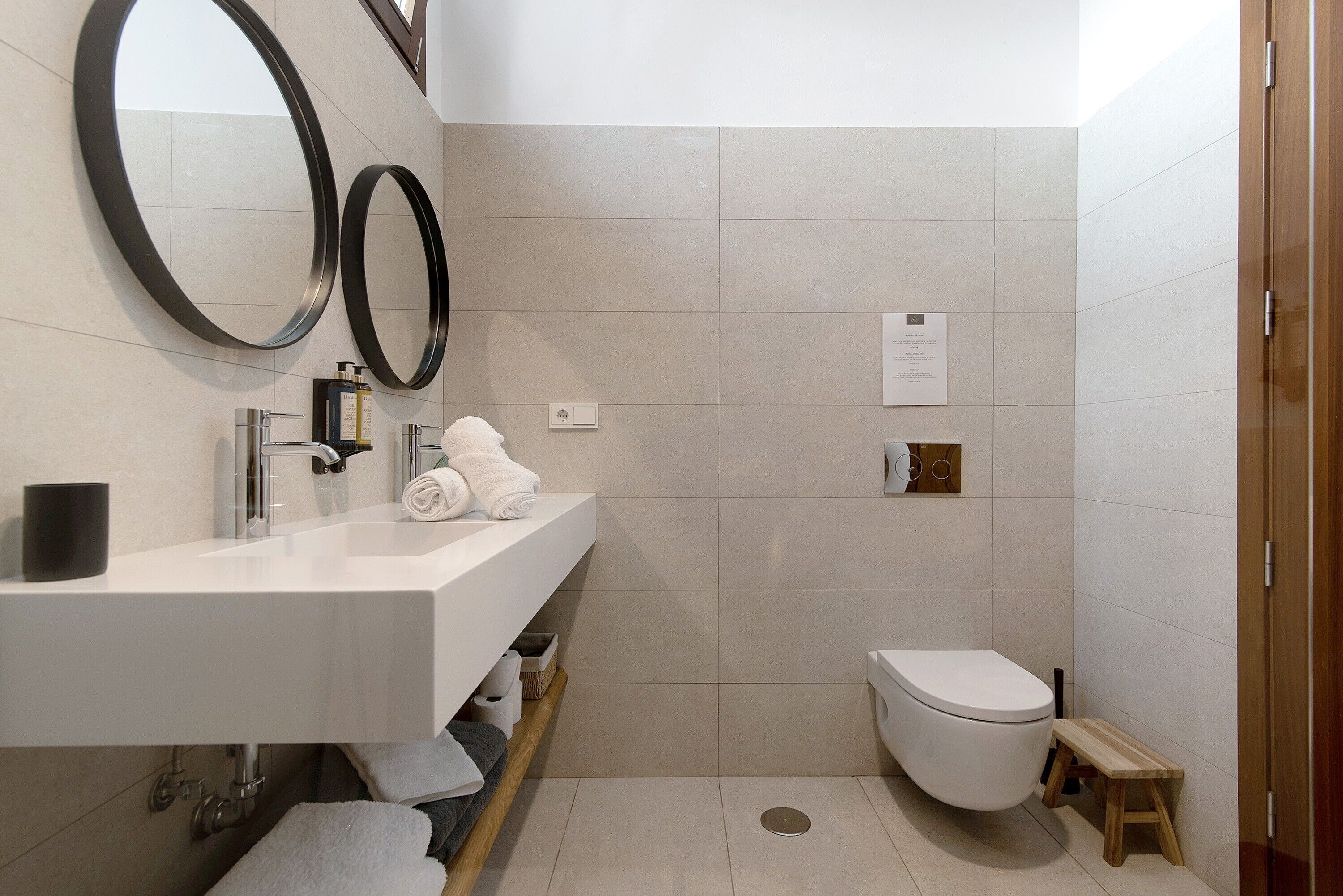 Un baño moderno con un lavamanos blanco, dos espejos, un inodoro y un taburete de madera.