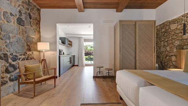 A bedroom with a wooden bed, a bench, a lamp, a stone wall, and a view into the kitchen. - Country House 'Iris - Finca La Suerte Grande' with Mountain Views and Wi-Fi (Telde)