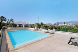 A swimming pool with lounge chairs, a covered area, and a view of distant buildings and hills.