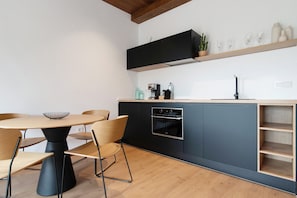 A modern kitchen with a round dining table, chairs, a built-in oven, and a wooden shelf above the counter.
