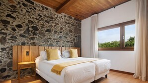 A bedroom with a stone wall, wooden ceiling, a large bed with white bedding, a nightstand, and a window with curtains. - Holiday Home 'Musa - Finca La Suerte Grande' with Mountain Views and Private Terrace (Telde)