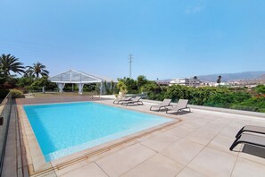 A swimming pool with lounge chairs, a covered area, and a view of distant buildings and hills.