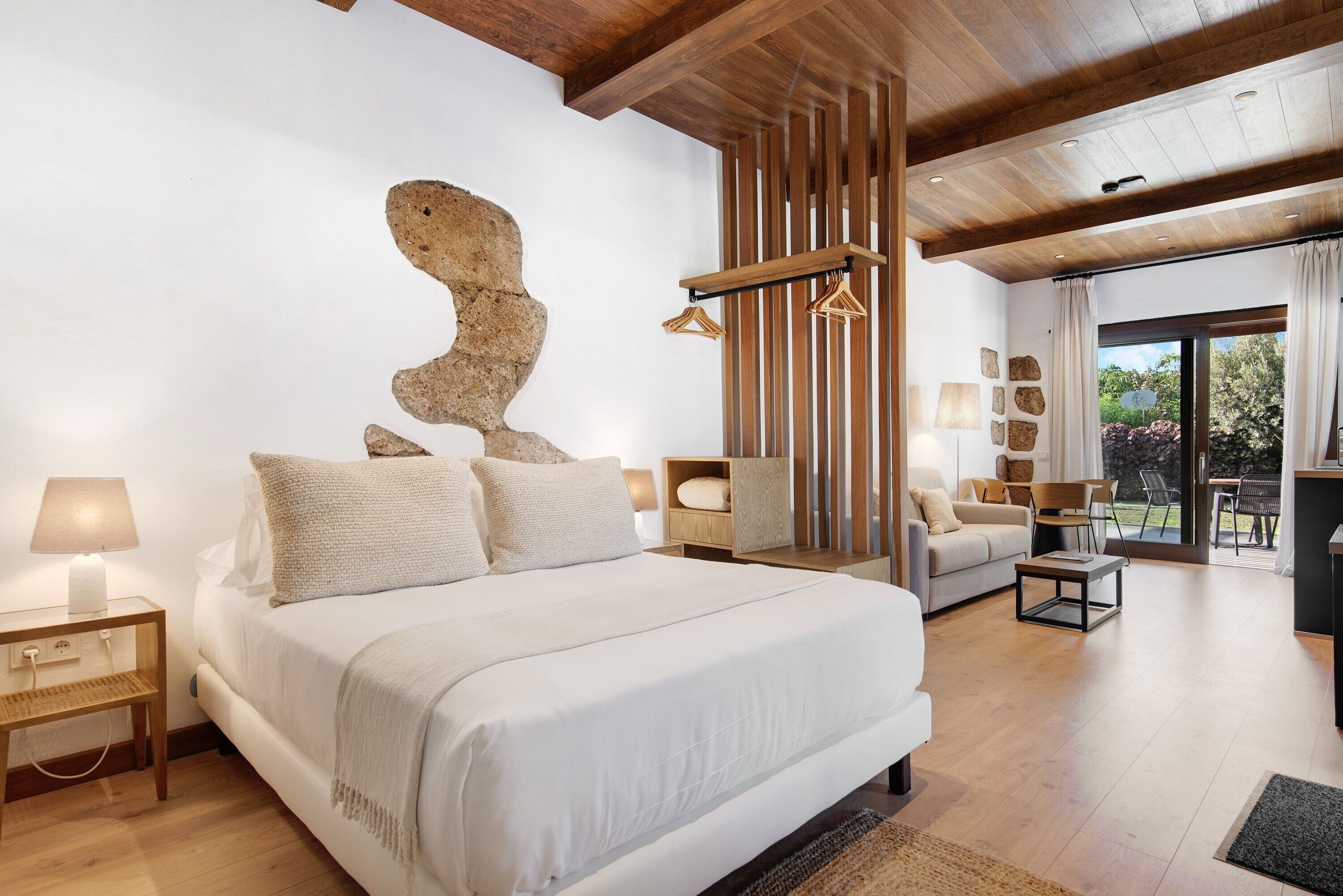 Une chambre moderne avec un grand lit, des poutres en bois, un mur en pierre et une vue sur l’extérieur.