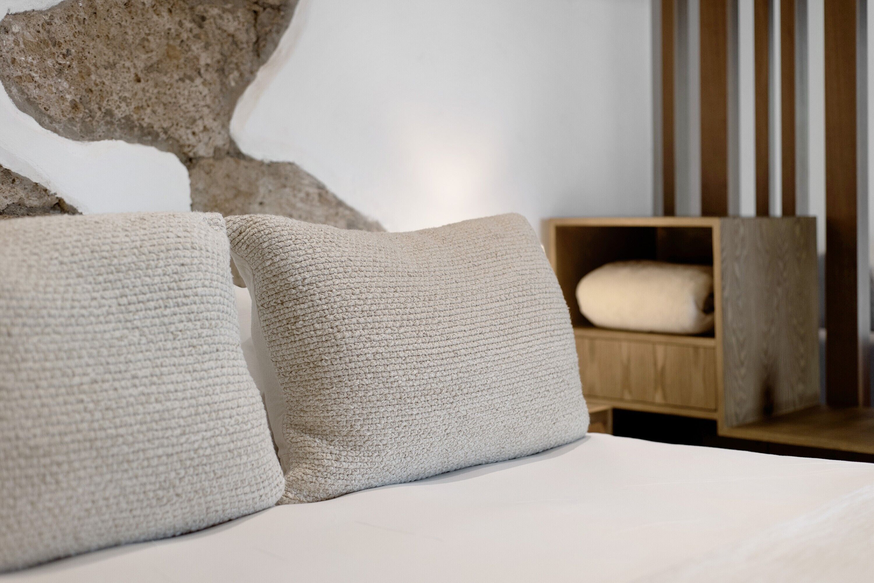 Una cama con dos almohadas texturizadas, una mesita de noche de madera con un almohadón blanco y una pared de piedra al fondo.