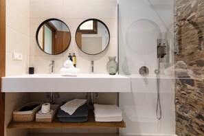 Een badkamer met een witte wastafel, twee ronde spiegels, een douche en een stenen muur.