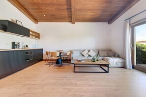 A modern kitchen with a built-in oven, a dining area with a wooden table and chairs, a sofa with cushions, and a large window with curtains.