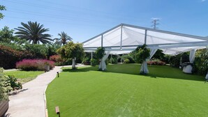 Une pelouse bien entretenue, surmontée d’une structure blanche en forme de tonnelle, et entourée de diverses plantes et arbres.