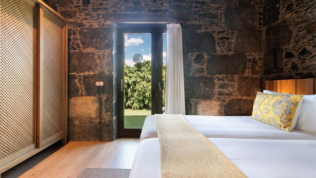 Une chambre avec un mur en pierre, un lit recouvert de linge de maison blanc, un plancher en bois et une vue sur la verdure à l’extérieur.