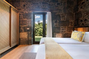 A bedroom with a stone wall, a bed with white bedding, a wooden floor, and a view of greenery outside.