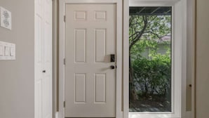 Une porte blanche au design moderne, une porte vitrée donnant sur un jardin avec des arbres et des arbustes, et un petit tapis sur le sol.