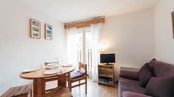 Una stanza con un tavolo da pranzo in legno, sedie, un divano viola, una TV su un mobile e una finestra con tende.