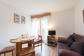 Una habitación con una mesa de comedor de madera, sillas, un sofá púrpura, un televisor sobre un mueble bajo y una ventana con cortinas.