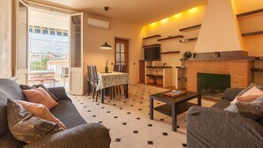 Un salon comprenant un canapé, une table basse, une cheminée, un coin repas avec une table et des chaises, et une vue sur l’extérieur par une porte ouverte.