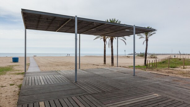 Une terrasse en bois qui mène à une plage, avec un espace abrité, des palmiers et une plage de sable.