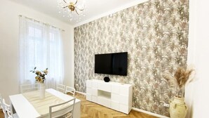 Een moderne woonkamer met een houten visgraatvloer, een witte eettafel met stoelen, een witte console en een flatscreen televisie die aan een wand met bloemenbehang is bevestigd.